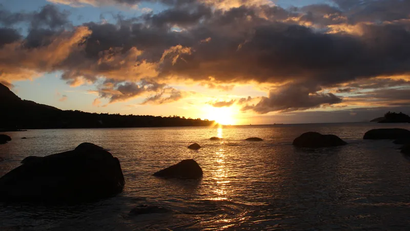 Pôr do sol na Praia do Julião, em Ilhabela: sol baixando atrás do morro, nuvens douradas e pedras escuras na água do canal