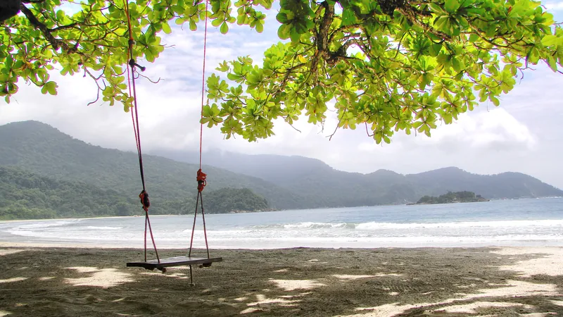 Balanço de corda pendurado numa amendoeira na areia da Praia de Castelhanos, em Ilhabela, com o mar e as montanhas da baía ao fundo